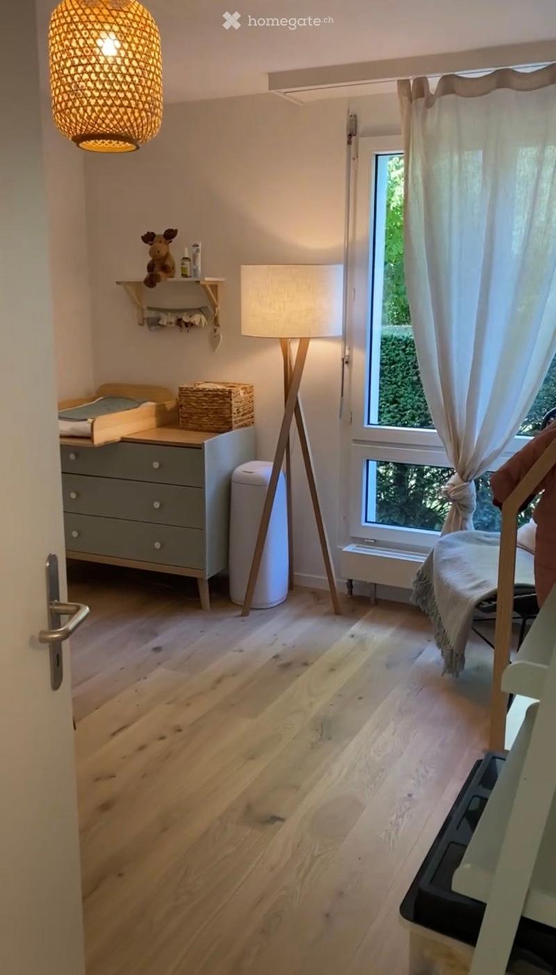 Cozy bedroom with wooden flooring, large window with white curtains, bed with white bedding, nightstand with lamp, drawer unit, wicker basket, and decorative shelf with a toy and small items.
