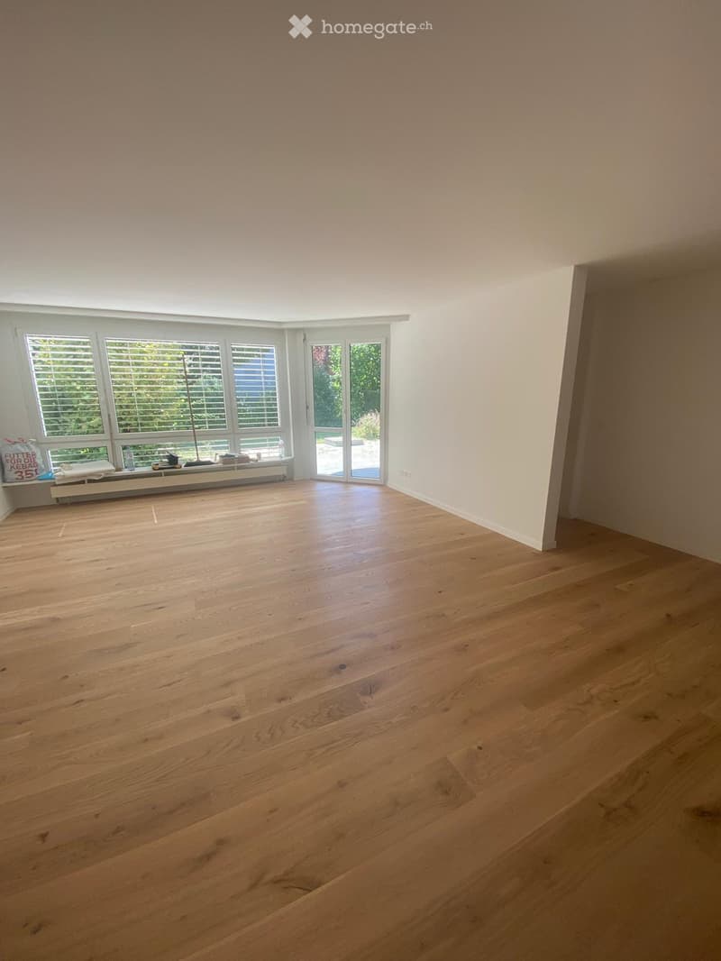 Empty room with light wooden flooring, large windows with blinds, and a sliding glass door leading outside.
