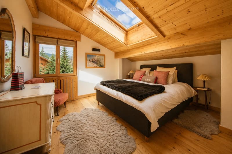Cozy bedroom with wooden beams, skylight, large bed with pillows and blankets, nightstands with lamps, a chair, a mirror, and a small table with a bag on it.