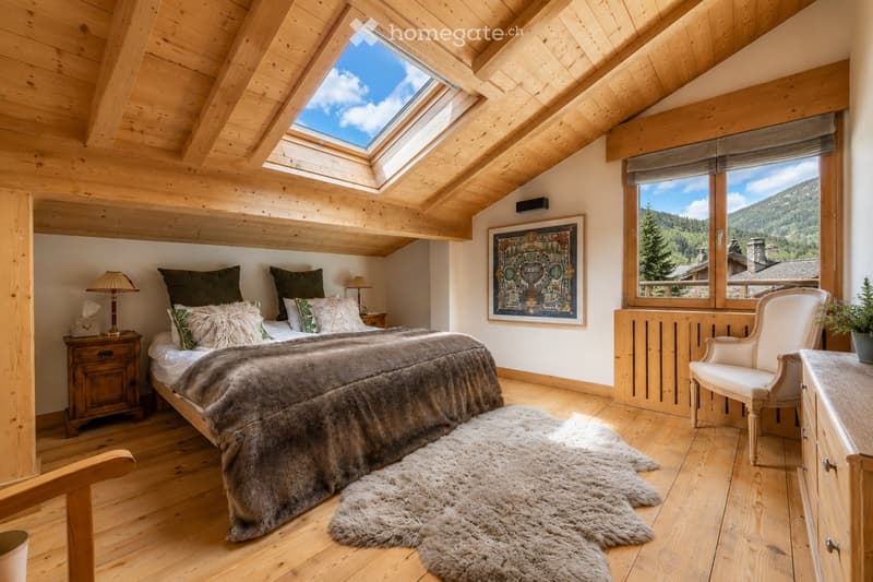 Cozy bedroom with wooden beams, skylight windows, large bed with fur throw, nightstand, armchair, and a scenic mountain view.