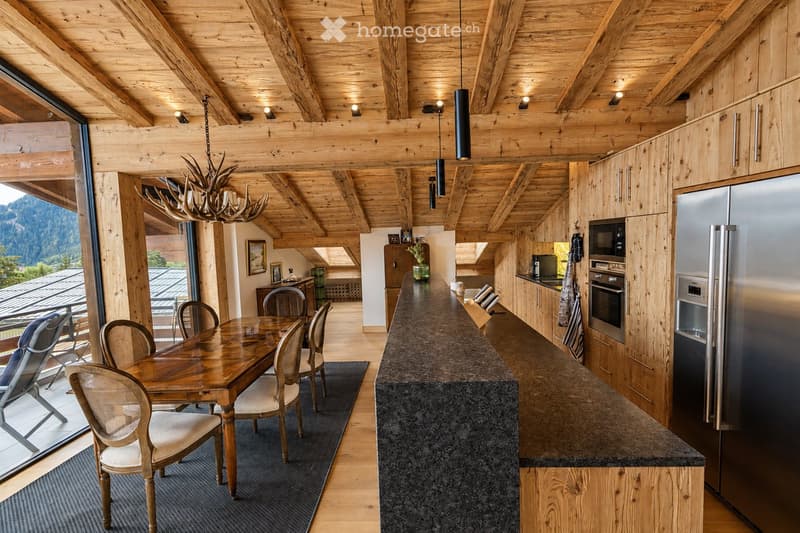 A modern, open-concept kitchen and dining area with exposed wooden beams, a large island, and a dining table with chairs. The space features stainless steel appliances, wooden cabinets, and a chandelier made of antlers.