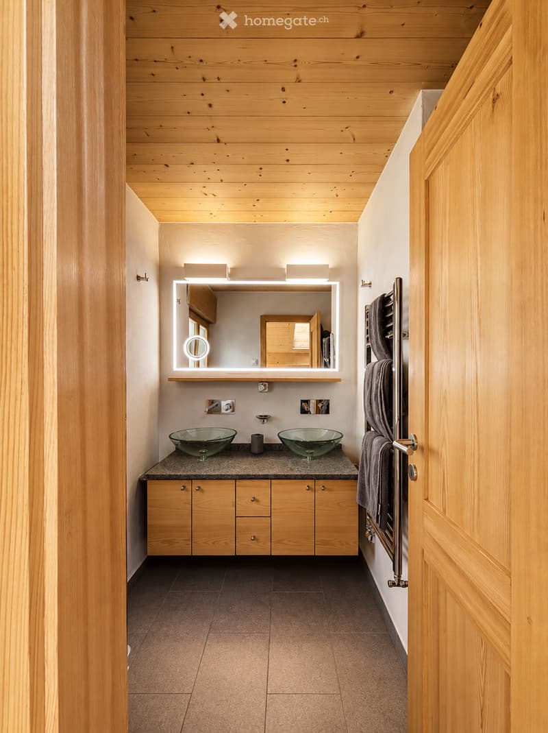 Modern bathroom with wooden walls and ceiling, two glass sinks, a large illuminated mirror, and a towel rack with a towel.