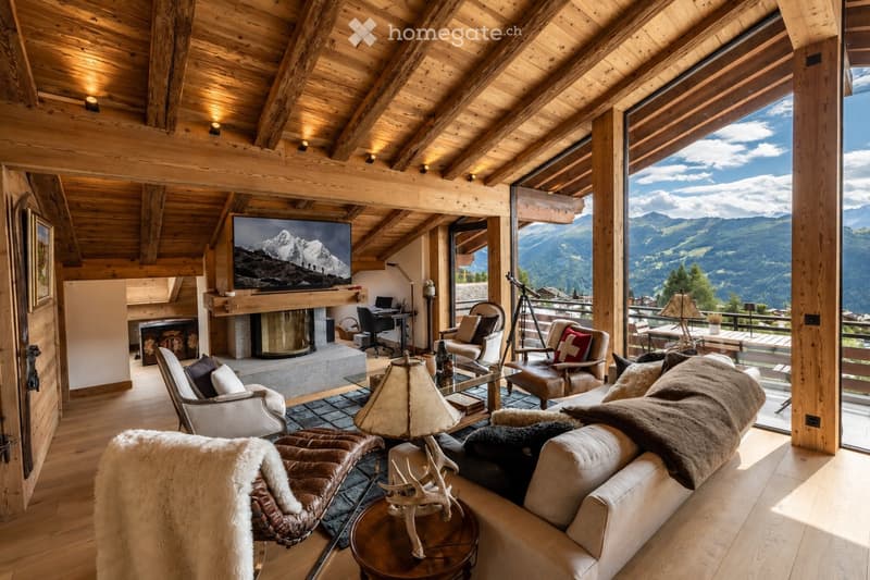 Cozy living room with exposed wooden beams, large windows offering mountain views, a fireplace, a sofa, armchairs, a coffee table, and a television.