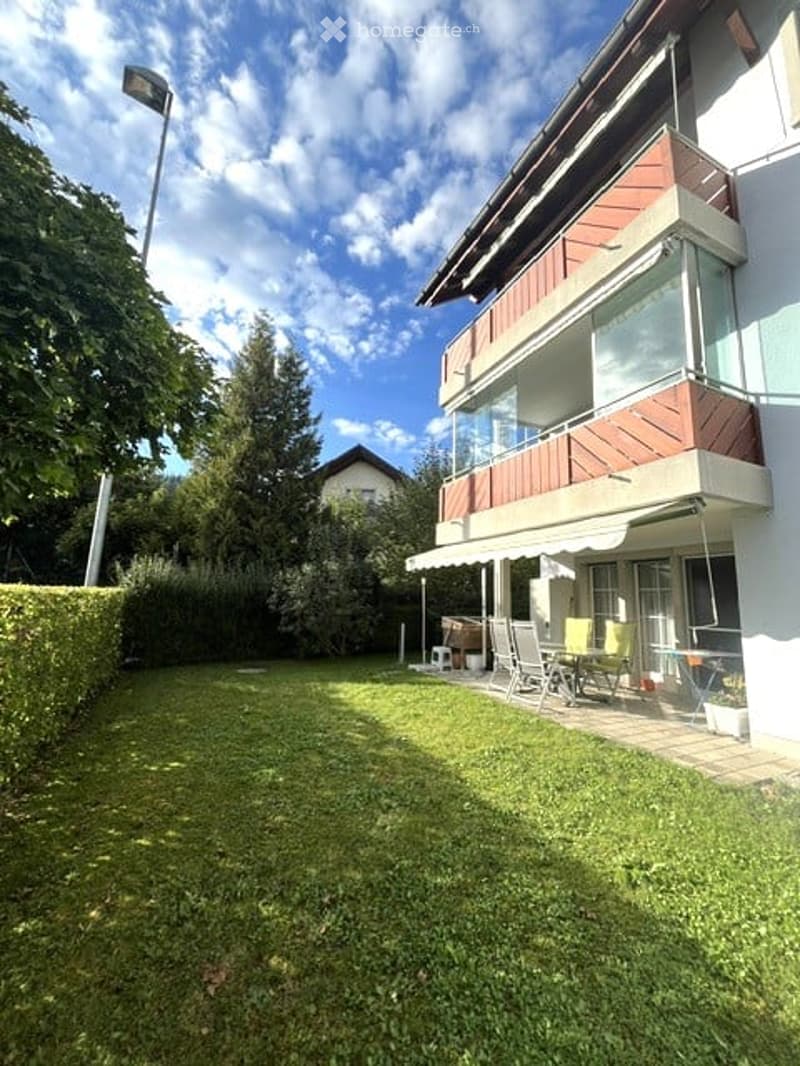 Well-maintained garden with a neatly trimmed hedge, a patio area with outdoor furniture, and a modern house with a balcony and large windows.