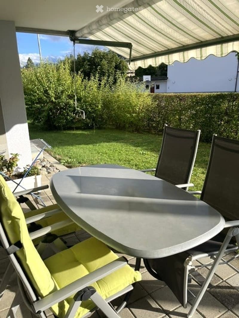 Balcony with a round table and four chairs, surrounded by greenery and a clear sky.