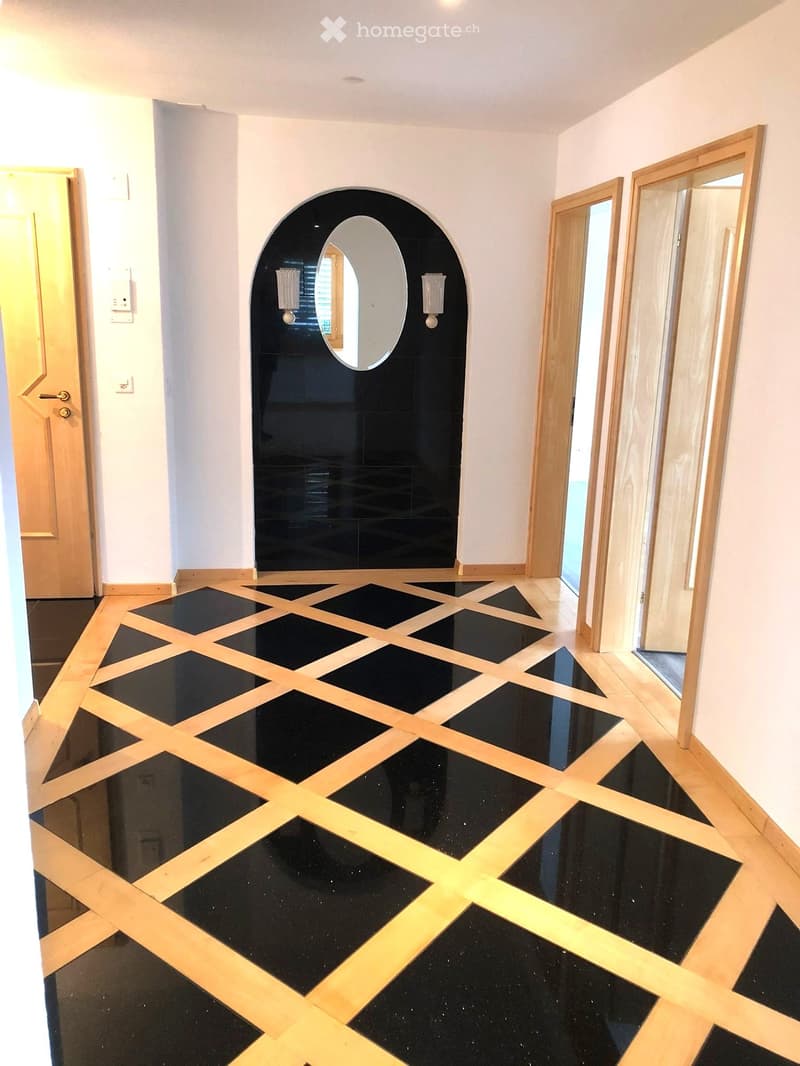 Hallway with black and light wood patterned flooring, white walls, and a black arched doorway with a round mirror and wall-mounted lights.