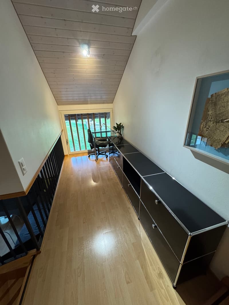 A narrow hallway with wooden flooring, white walls, and a slanted ceiling. A small black cabinet is on the right, and a glass door leads to a balcony area with a desk and chair visible beyond.