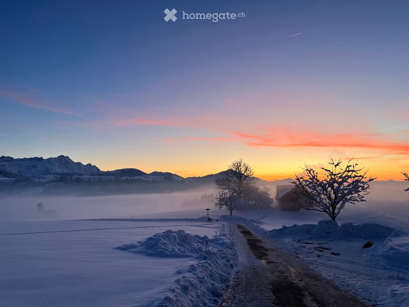 A serene winter landscape with snow-covered fields, a winding road, and distant mountains under a colorful sunset sky.
