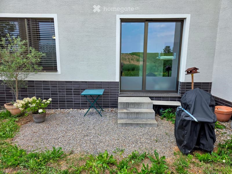 Garden area with a small table, potted plants, a covered grill, and a birdhouse near the entrance of a house.