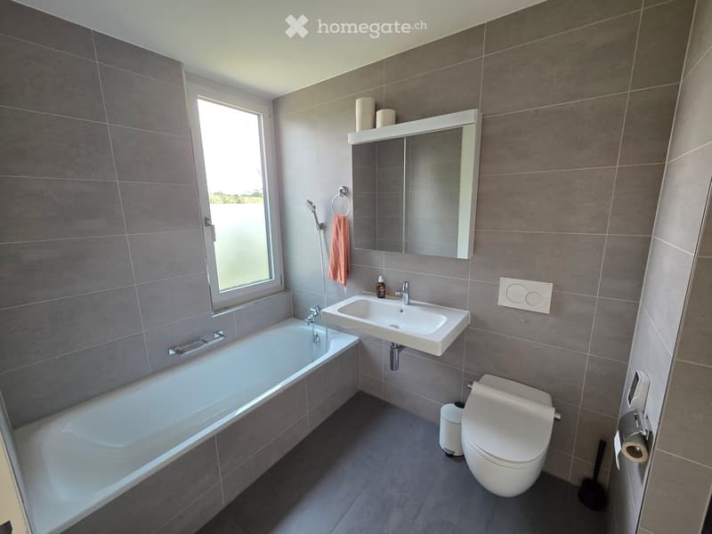 Modern bathroom with grey tiled walls, a bathtub, a sink, a toilet, a mirror, and a window providing natural light.