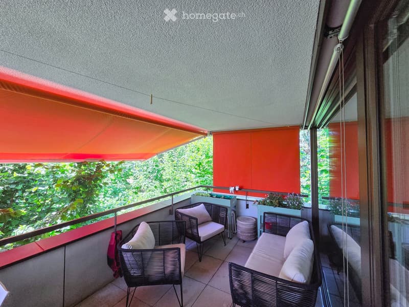 Balcony with wicker furniture, including a sofa, chairs, and a small table, with a view of greenery and a retractable awning.