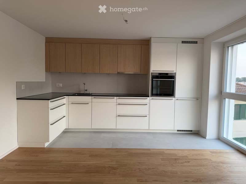 Modern kitchen with light wood cabinets, white lower cabinets, stainless steel sink, and built-in oven. The space features a large window and wooden flooring.