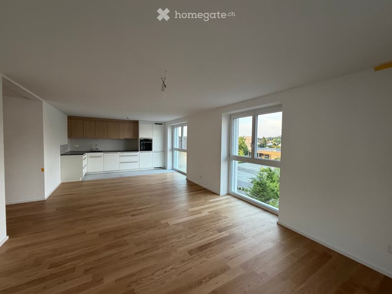 Open-plan unfurnished room with wooden flooring, large windows, and a small kitchen area with cabinets and appliances.