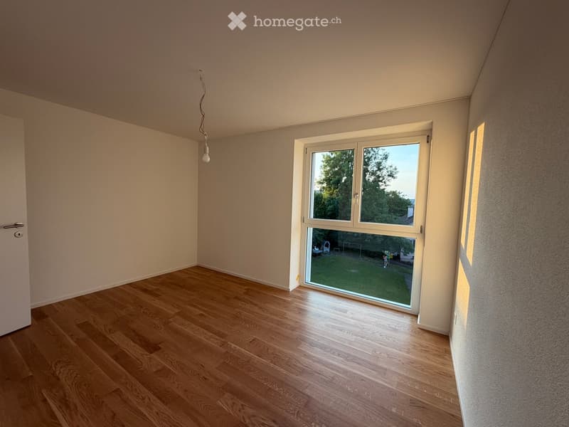 Empty room with wooden flooring, white walls, and a large window overlooking a green yard.
