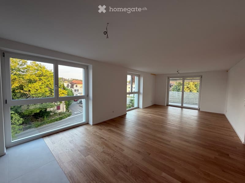 Empty room with wooden flooring, large windows, and sliding doors leading to a balcony or outdoor area.