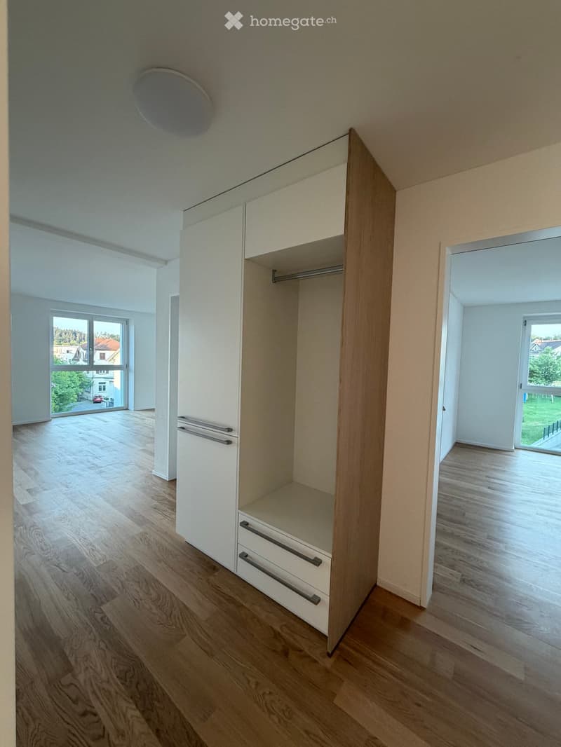 Empty room with wooden flooring, white walls, and built-in closet with drawers and shelves. Large windows provide natural light and views of the outdoors.