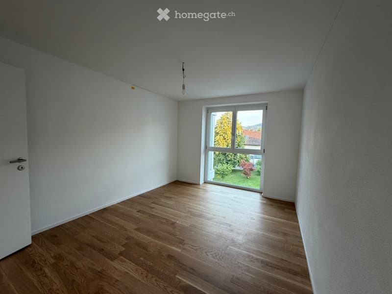 Empty room with light-colored walls, wooden flooring, and a large window showing a view of greenery outside.