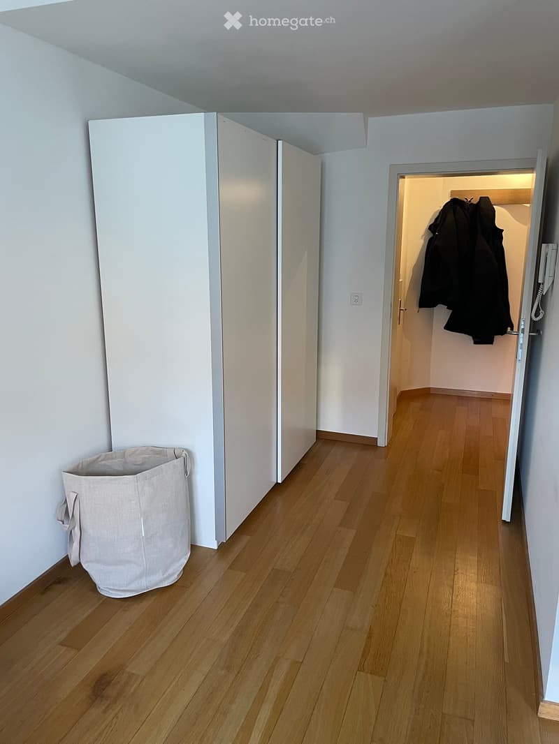 Empty hallway with light wooden flooring, white walls, and a partially open door leading to a closet area with coats hanging inside.
