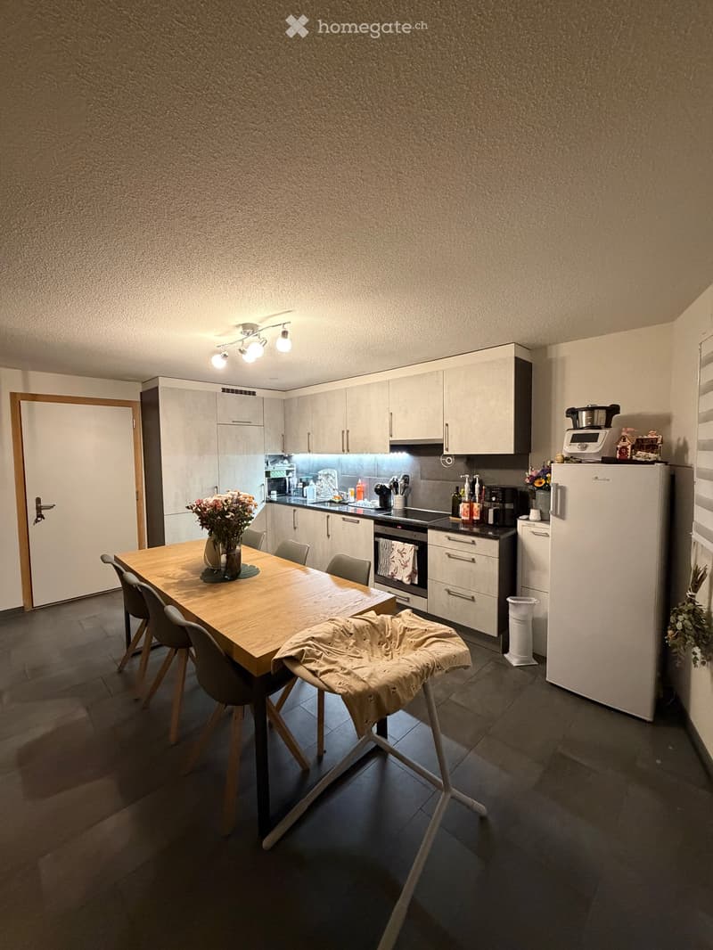 Modern kitchen with light grey cabinets, a wooden dining table with four chairs, a refrigerator, oven, microwave, coffee maker, and a flower vase on the table.