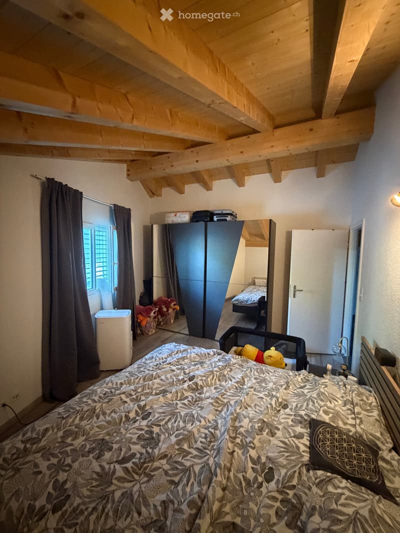 A cozy bedroom with a large bed, patterned duvet, and a mirror reflecting part of the room. The space features exposed wooden beams on the ceiling, a window with dark curtains, a wardrobe, and a small air conditioner unit near the window. Toys and a baske