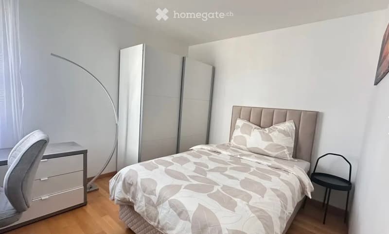 Modern bedroom with a large bed, grey and white bedding, a sleek wardrobe, a desk with a chair, and a floor lamp.