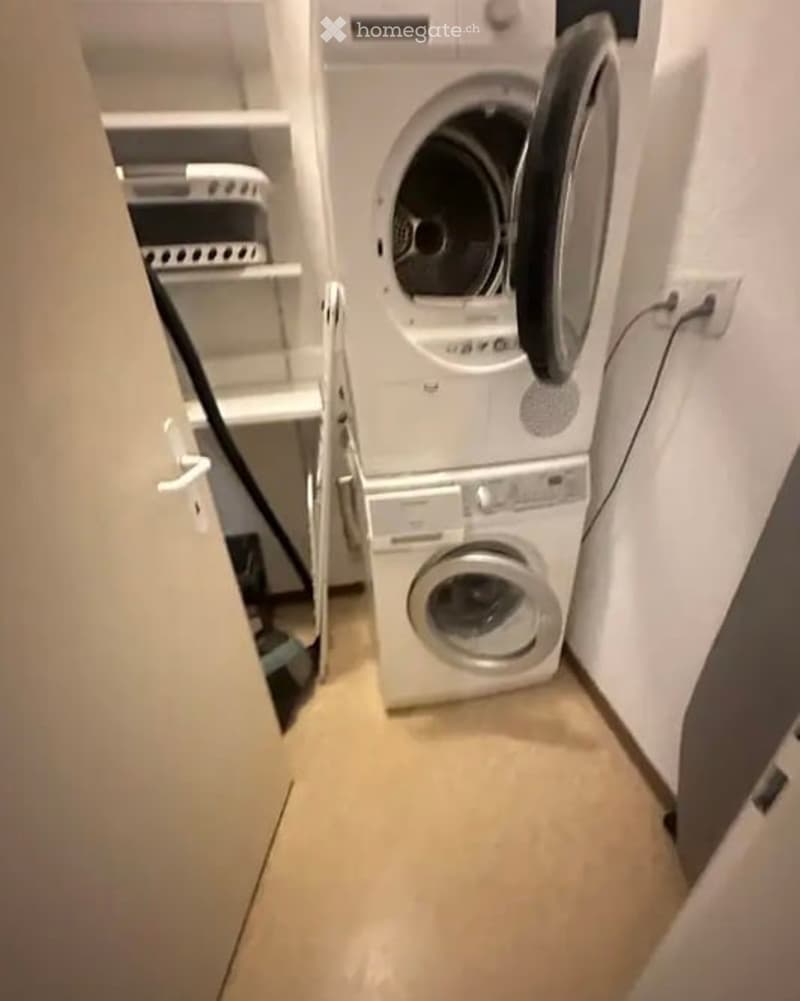 Compact bathroom with a stacked washing machine and dryer unit, a small shelf with folded towels, and a tiled floor.