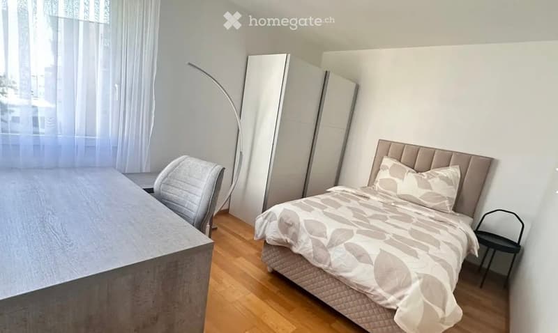Modern bedroom with a large bed, a wardrobe, a chair, a table, and a floor lamp. The room features wooden flooring and large windows with sheer curtains.