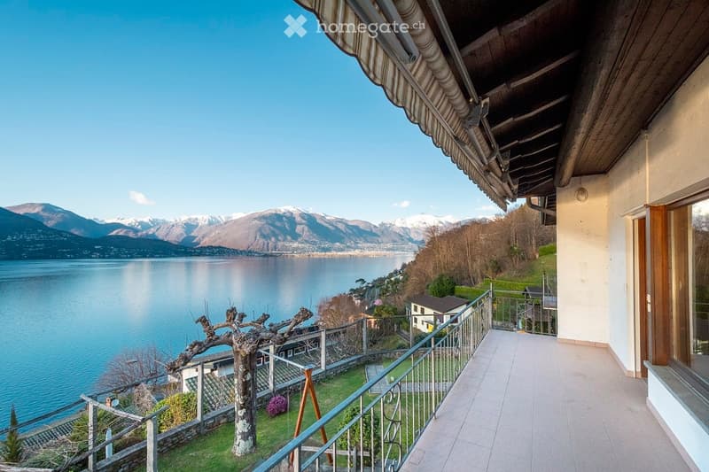 Balcony overlooking a serene lake with distant snow-capped mountains, featuring a tiled floor, metal railing, and a wooden awning.