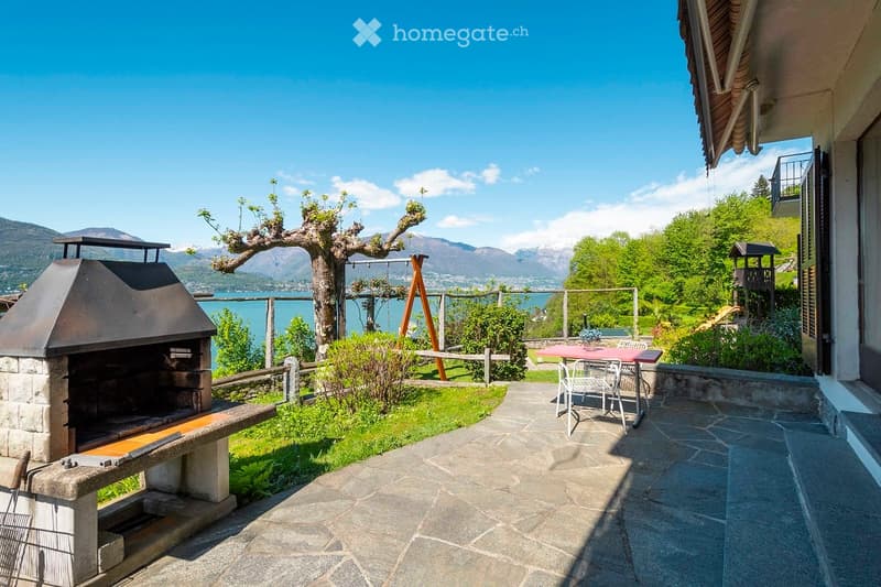 A serene outdoor garden area with a stone barbecue, a dining table with chairs, a swing, and a scenic view of a lake and mountains.