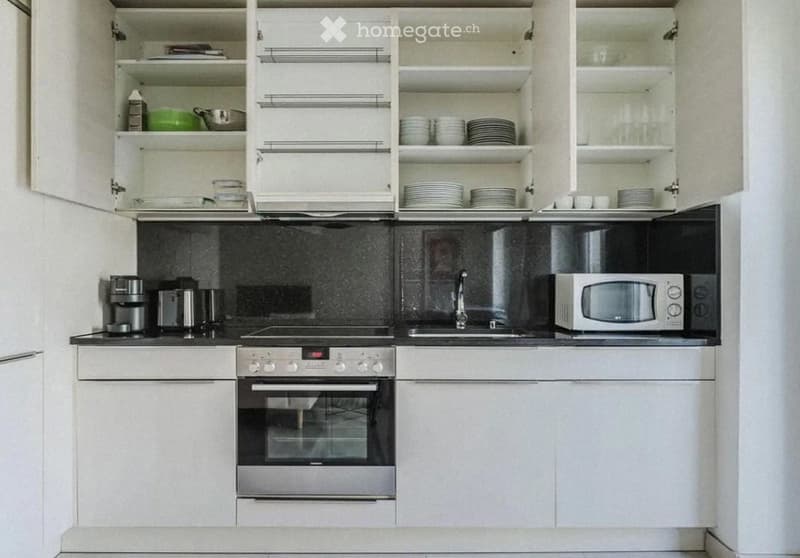 Modern kitchen with white cabinets, stainless steel appliances, and a black backsplash. Visible items include an oven, microwave, coffee maker, and various dishes in the open cabinets.