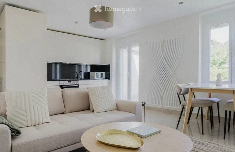Modern, open-plan living and dining area with a light-colored sofa, coffee table, dining table with chairs, and a sleek kitchen area featuring a microwave and oven. The space is bright with large windows and a decorative wall design.