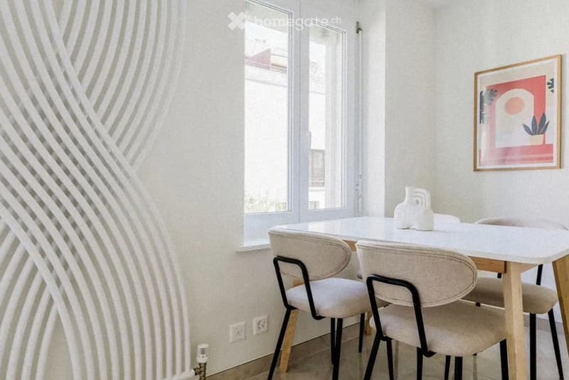 Modern dining area with a white dining table, four beige chairs, and decorative vases. Large windows provide natural light, and a framed abstract artwork adorns the wall.