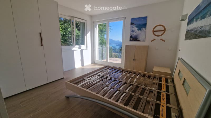 Bright bedroom with wooden floor, large windows, and a view of the lake and mountains. Features a modern bed frame and a built-in wardrobe.