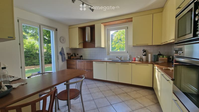 Bright kitchen with light yellow cabinets, wooden countertops, and tiled flooring. Features a dining table with chairs, microwave, oven, sink, and various kitchen utensils.