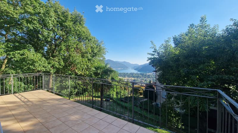 Balcony with tiled floor, metal railing, overlooking a scenic landscape with trees, mountains, and a lake.