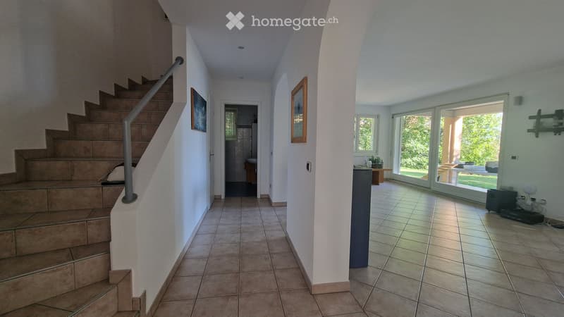 Bright hallway with tiled floor, staircase leading upstairs, and open doorway to a room with large windows and outdoor view.