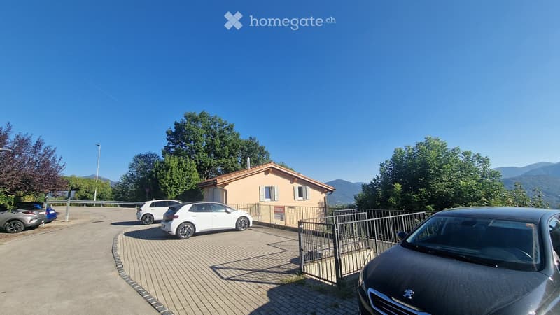 A small, single-story house with a tiled roof and white shutters, surrounded by trees and mountains, with a paved parking area and several parked cars.