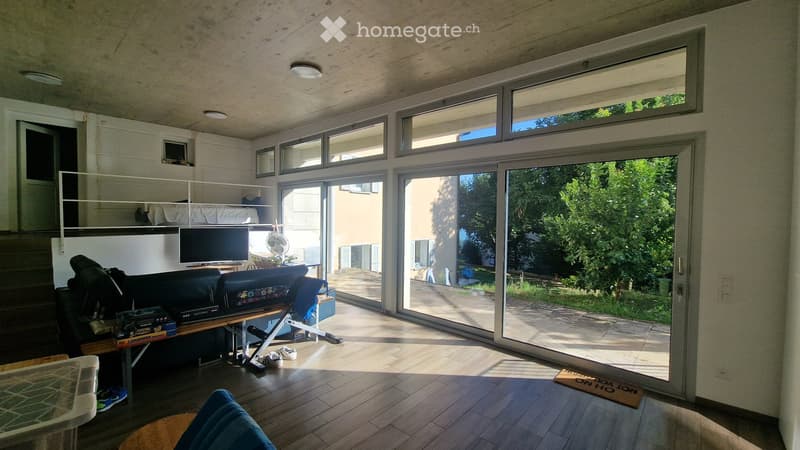 Modern living room with large windows and a sliding glass door leading to a balcony. The room features a sofa, a small table, a screen, and a fan. The floor is wooden, and the ceiling has recessed lighting.