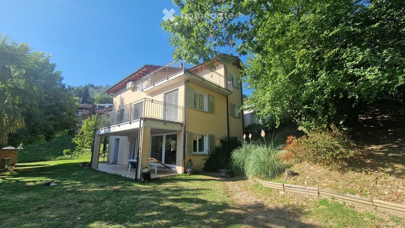 Two-story yellow house with balconies, surrounded by greenery and a well-maintained lawn.