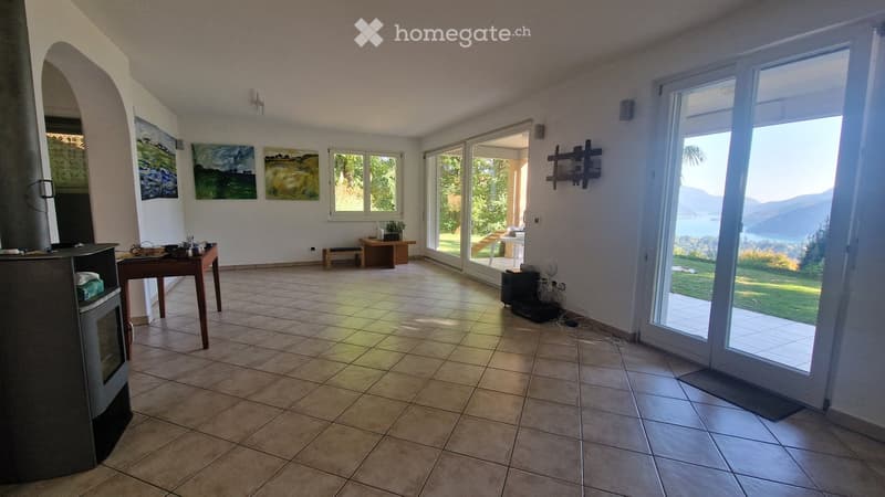 Bright living room with tiled floor, large windows, and a scenic view of a lake and mountains. Features include a fireplace, a small table, a bench, and wall-mounted shelves with artwork.