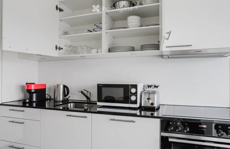 Modern kitchen with white cabinets, black countertops, and stainless steel appliances including a coffee maker, kettle, microwave, toaster, and oven. Open shelving displays plates and glasses.
