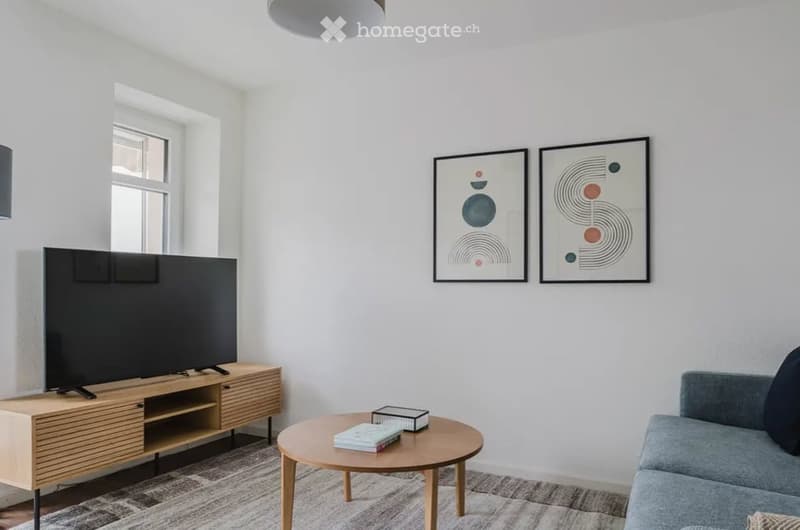 Modern living room with white walls, grey sofa, wooden coffee table, TV stand, and two abstract art pieces on the wall.