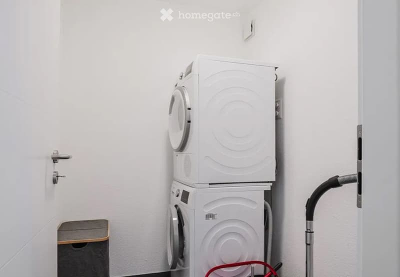 A small, white-walled room with a stacked washing machine and dryer unit, a vacuum cleaner, and a small wooden shelf.