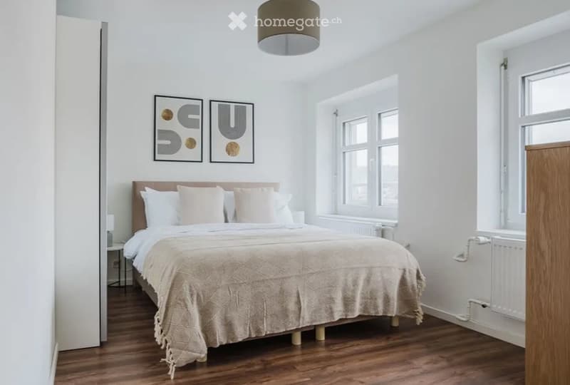 Bright bedroom with wooden flooring, large bed with pillows and blanket, nightstand with lamp, radiator, and two picture frames on the wall.