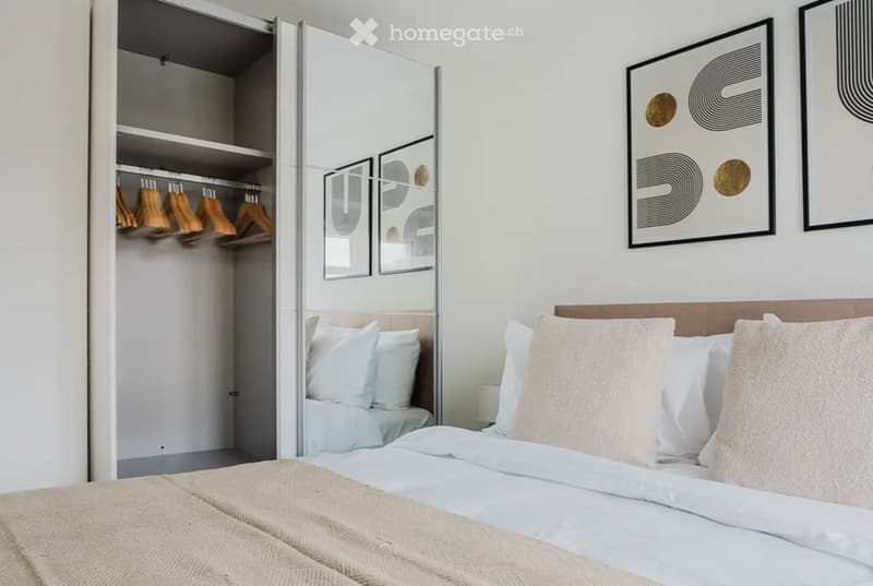 Modern bedroom with a neatly made bed, white pillows, a wardrobe with hanging clothes, and framed artwork on the wall.