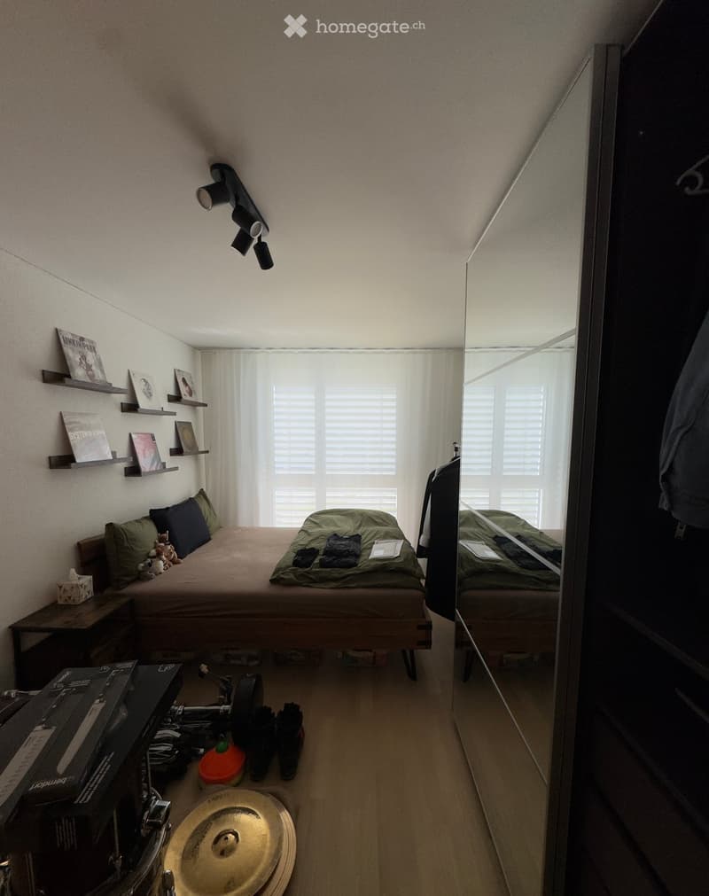 Cozy bedroom with a wooden bed, green bedding, nightstand, shelves with books and picture frames, a large mirror, and various items including a drum and guitar.