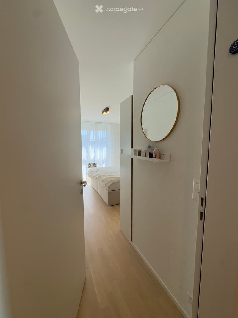 A modern hallway with light wooden flooring, white walls, and a round mirror on the wall. A small shelf holds various items, and a bedroom is visible at the end of the hallway.