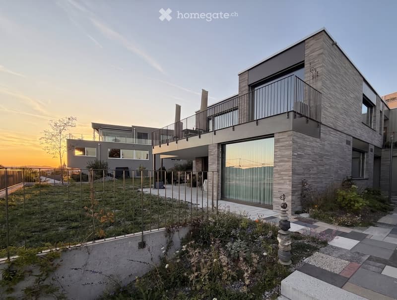 Modern two-story house with grey brick facade, large windows, and a balcony on the upper level. The surrounding area includes a landscaped garden with a stone pathway and a small fence.