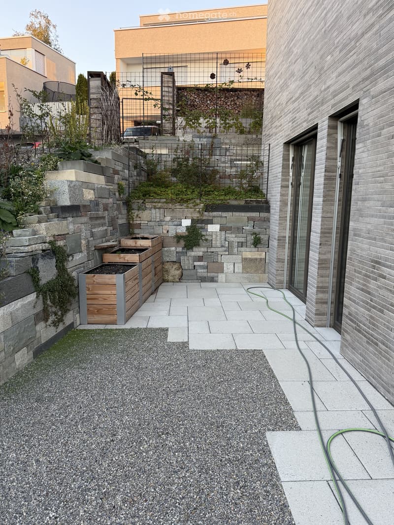 Modern outdoor space with stone and gravel surfaces, wooden bench, greenery, and a raised garden bed.