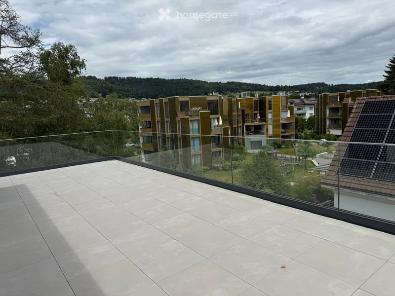 Modern rooftop terrace with large glass railing, tiled floor, and scenic view of surrounding buildings and greenery.